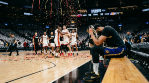 NBA playoff scene showing a dejected player on the bench after a first-round elimination loss while opposing team celebrates